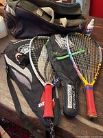 Photo showing four racquets, two with racquet covers, along with a carrying bag and spray bottle of glasses cleaner on a wooden surface.