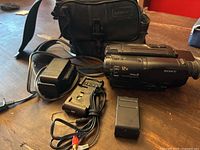 Full view of Sony camcorder with charger, batteries, battery discharger, and black carrying bag on wooden surface.
