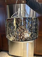 Chandelier being held up in front of kitchen appliances showing stainless steel frame with geometric pattern.