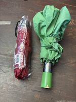 Two umbrellas placed side by side on a table, one green used umbrella open and fastened, one red ladies mini umbrella still in plastic packaging.