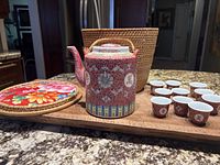 Front view of the vintage Chinese porcelain red patterned teapot placed next to 10 small matching red and gold teacups with a woven wicker basket and floral fabric lid in background.