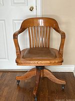 Front view of the antique wooden rolling chair showing vertical slat backrest, curved armrests, and brown leather seat cushion.