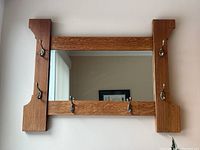 Full view of the wooden framed mirror mounted on the wall showing six metal coat hooks attached, three along the bottom edge and one on each side panel of the vertical wood frame.