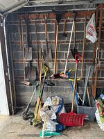 Full view of garden tools leaning against garage wall showing shovels, rakes, brooms, weed whackers and other tools.
