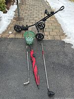 PowerBuilt golf push cart with three wheels, green basket containing golf balls, left-handed driver, right-handed chipper, and red and black umbrella laid out on pavement with snow in background.