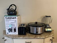 Photo showing all kitchen appliances in the lot: Cuisinart coffee maker in box, black kettle, black toaster, stainless steel slow cooker, and vintage Pulse Matic blender.