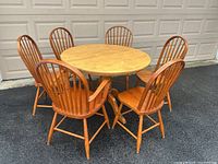 Full view of maple round dining table surrounded by four straight-back chairs and two armchairs in light honey finish outside in front of garage doors.