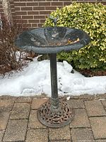 Full view of metal birdbath on paved outdoor surface with shrub background showing overall condition and details