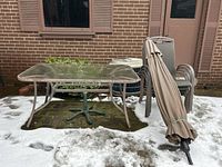 Outdoor patio furniture set showing rectangular glass top table, stacked chairs, and folded umbrella with iron base on a snowy ground outside near building wall.