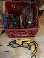 Red plastic crate filled with assorted hand tools and yellow DeWalt plug-in drill lying in front of it on the floor.