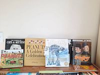 Six hardcover books standing upright on a wooden surface, showing spines and front covers with titles Peanuts, Star Wars, Cats & Kittens and more.