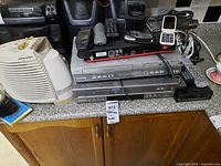 Photo of various electronics on countertop including Panasonic stereo, Samsung DVD VCR player, Black & Decker heater, remotes, cords, and cell phones.