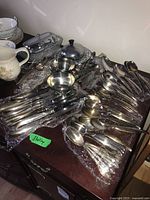 Overview of assortment of flatware including spoons, forks, and a metal butter dish with lid on a wooden surface with bubble wrap beneath.