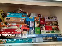 Wide angle of stacked boxed board games and jigsaw puzzles including identifiable titles and piece counts.