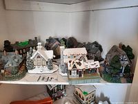 Photo showing 7 miniature houses with snow-covered roofs, trees, and village details on a shelf.
