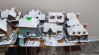 Overview of six Thomas Kinkade Hawthorne Village buildings with snow on roofs and surrounding pine trees, grouped on a table.