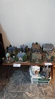 Photo of 12 Thomas Kinkade Hawthorne Village miniature houses arranged on a table and boxes. Various styles and details visible including a gazebo and multiple homes.