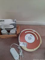 iRobot Roomba vacuum robotic cleaner in reddish-brown and cream color, positioned next to a charging dock on carpeted floor with a tag on it.