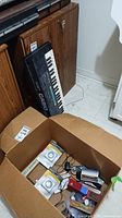 Photo showing Casio MT-540 keyboard leaning upright against wood furniture and a large brown cardboard box containing indoor basic timer units and various small items.