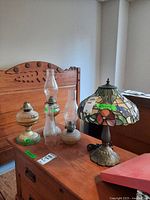 Tiffany style lamp with stained glass floral shade and three vintage oil lamps with glass chimneys on a wooden table in front of a wooden headboard.