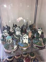 Photo showing eleven Thomas Kinkade music boxes displayed inside a glass cabinet. The boxes depict miniature garden scenes with gazebos, flowers, pathways, and churches atop round bases.