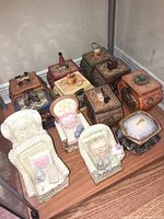 Photo of 12 heirloom porcelain music boxes with varied shapes including small furniture and rustic cottage motifs, arranged on a wooden floor in a glass display case.