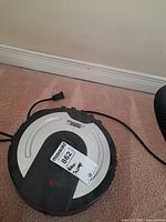 Top view of Robo Sweep robotic vacuum cleaner showing circular design with black and silver colors, placed on carpeted floor with power cable attached.