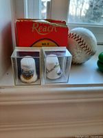 Two ceramic thimbles displayed in clear cases with a red cardboard box and a baseball behind