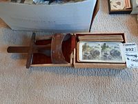 Photo of wooden stereoscope viewer with handle, stereograph cards, and storage box on carpet.