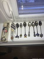 Lot displayed on window sill showing all 9 souvenir spoons, one in packaging and others with various detailed handles and emblems.