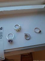 Top view of four rings placed on a white surface; includes purple gemstone ring, yellow gemstone ring, floral dark gemstone ring, and souvenir style ring with blue stone.
