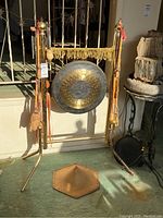 Frontal view of brass-coated gong on metal stand with tassel fringe and hanging mallets, hat on floor