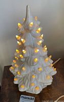 Front angle of the white ceramic Christmas tree ornament illuminated with warm yellow lights, showing tree shape and light bulb details.