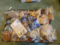 Top view of assorted resealable bags containing small crafting seed beads, pearl-like beads, and wooden beads scattered in a cardboard box on a green carpet.