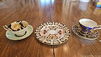 Three pieces on a wooden table: Aynsley teacup and saucer with floral pattern, Paragon teacup and saucer in cobalt blue with gold, decorative plate with geometric floral design.
