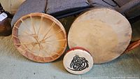 Front view of three drums: two natural hide drums and one small red-painted drum with black design