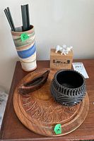 Photo showing the ceramic vase with earth tone stripes holding black rods, dark ceramic bowl, small carved wooden canoe, circular carved wood thunderbird plaque, and star marble stone bear carvings on wooden box.