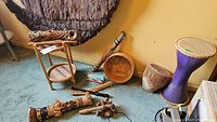 Wide shot showing two drums (purple hourglass drum and smaller natural wood drum), rainstick, nut shell shaker, carved wooden percussion instruments, wooden bowl, and a wooden stand.