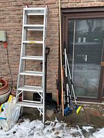 Full view of aluminum extension ladder leaning against brick wall with outdoor gardening and cleaning tools nearby.