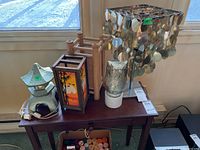 Five table lamps on wooden surface against window showing different styles and materials including metal, ceramic, wood, and glass. Includes visible cords and various heights.