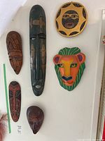 Photo showing six masks on a wall: a tall dark wood mask with carvings, a brown wood mask with patterns, a red-black bird-like mask, a lion mask in bright colors, a round sun mask, and a small brown mask.