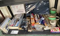 Photo showing a shelf with multiple office supplies including Trend pen box, stapler, DVD writer box, laptop bag, packs of charcoal pencils, masking tapes and various writing tools