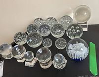 Overview of 15 clear glass paperweights of various bubble-filled and smooth designs with one floral accented piece.