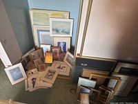 Wide view showing assorted framed and matted prints on the floor leaning against a wall, with various empty frames stacked beside them.
