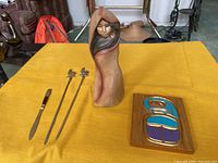 Full view of lot items on yellow surface including pottery figure, carved wood plaque, letter opener and two skewers