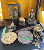 Multiple Thai Buddha figurines, stone trivets, incense holder, metal lotus wind chimes, and other decorative pieces arranged on a black surface.