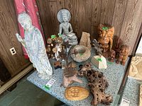 Photo showing various yard decor items including several Buddha statues, wood Tiki figures, metal flower sculptures, natural wood pieces, and a sun face decoration placed on a table against a wood-paneled wall.