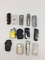 Full overhead view of all 13 assorted collectible butane lighters arranged on white surface showing various colors, designs, and shapes