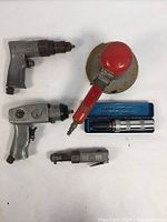 Five tools including air drill, dual-action sander, impact driver, and two pneumatic wrench ratchets displayed on white surface.