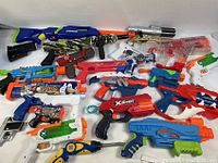 Top down overview of 17 assorted toy blasters in various colors and brands arranged on a white surface